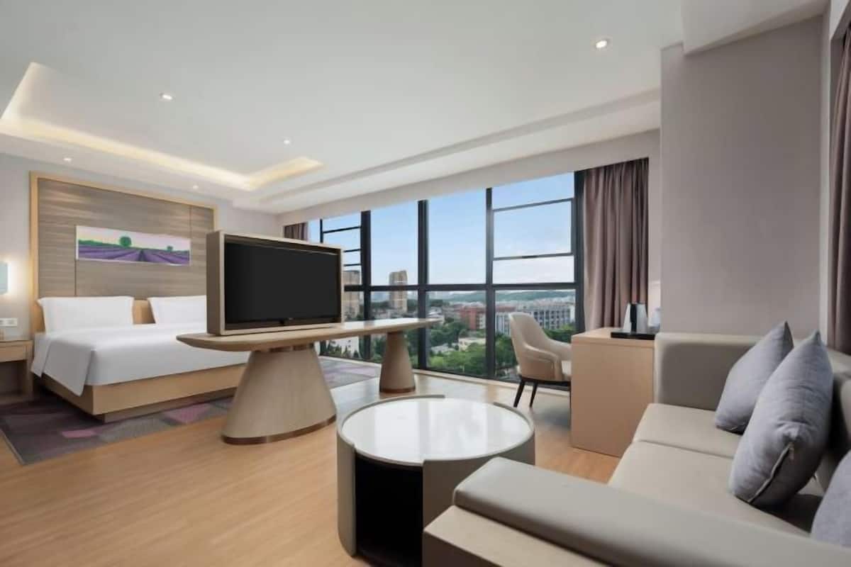 A modern hotel room with a bed, a TV on a stand, a sofa, a chair, and large windows offering a city view.