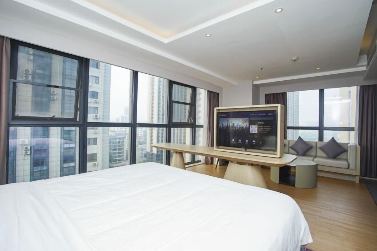 A modern hotel room with a large bed, a TV on a wooden stand, and large windows offering a city view.