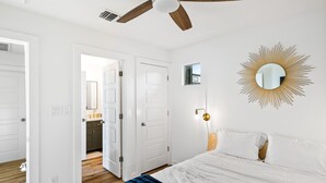 Une chambre à coucher comprenant un lit, un ventilateur de plafond, un miroir fixé au mur et une porte donnant sur une autre pièce.