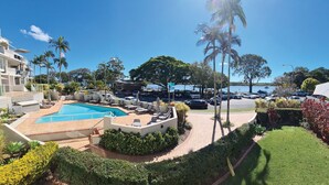 A pool area with lounge chairs and umbrellas, surrounded by greenery and a parking lot with cars. - Regatta Noosa - Top Floor Magic Views (Noosaville)