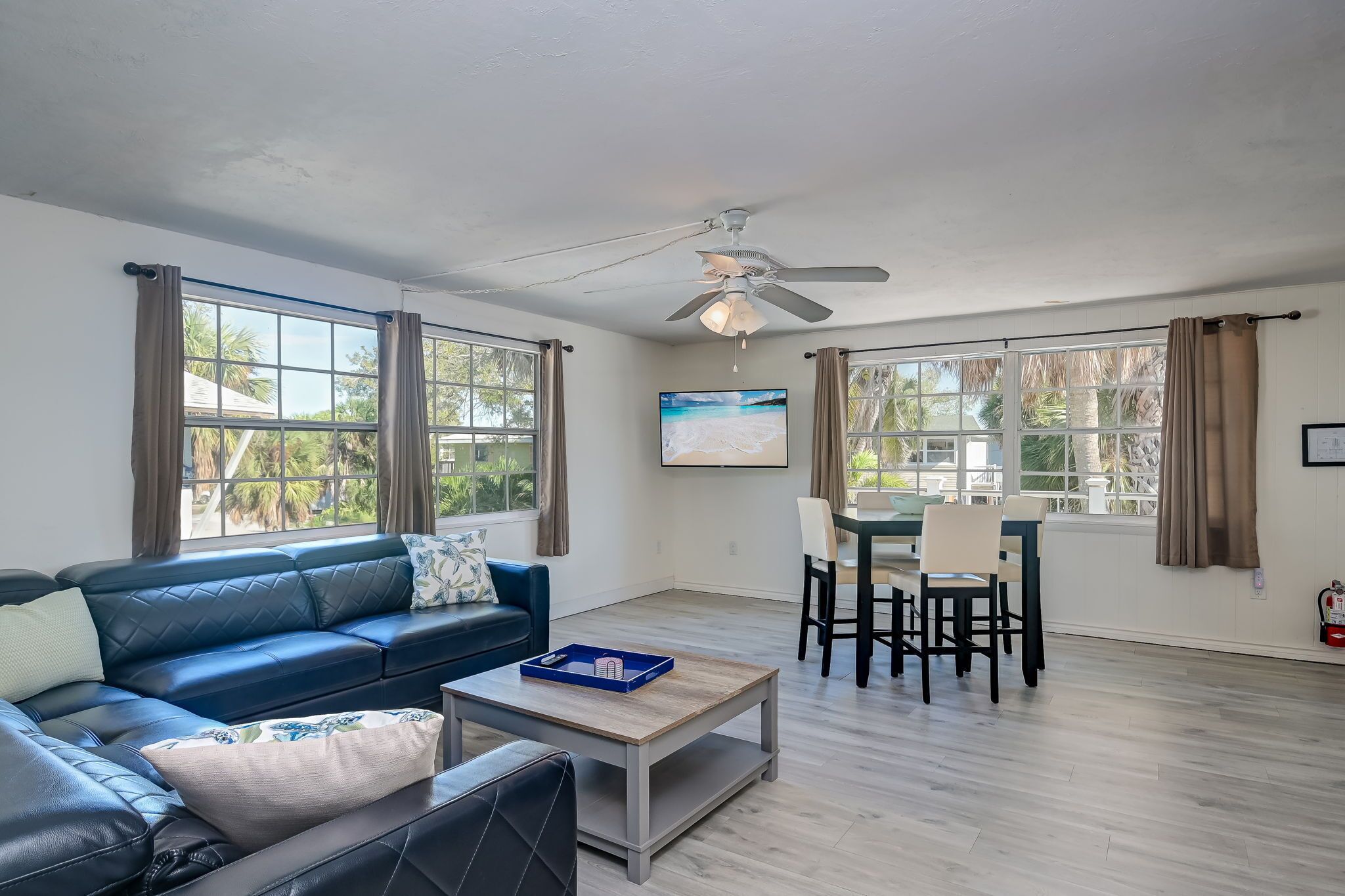 A living room with a blue sofa, a coffee table, a dining area with chairs, a ceiling fan, and large windows with curtains.