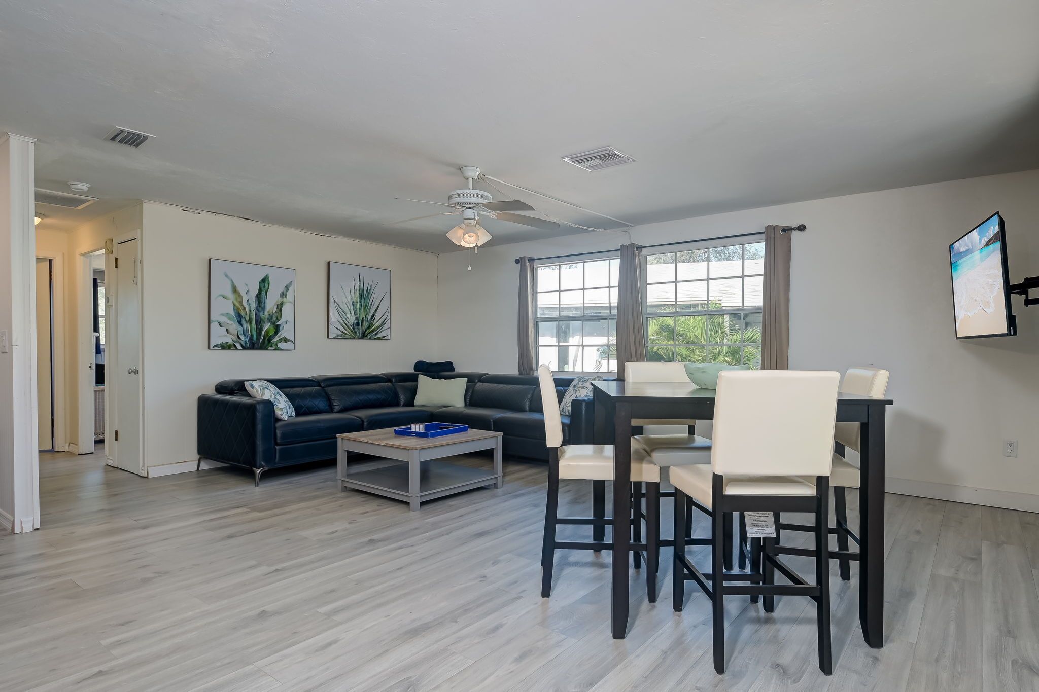 A modern living room with a dark-colored sofa, a coffee table, and a dining area with a table and chairs. There are large windows with curtains, a ceiling fan, and artwork on the walls.