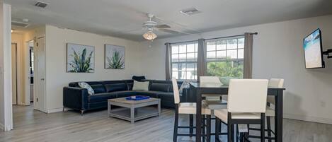 A modern living room with a dark-colored sofa, a coffee table, and a dining area with a table and chairs. There are large windows with curtains, a ceiling fan, and artwork on the walls.