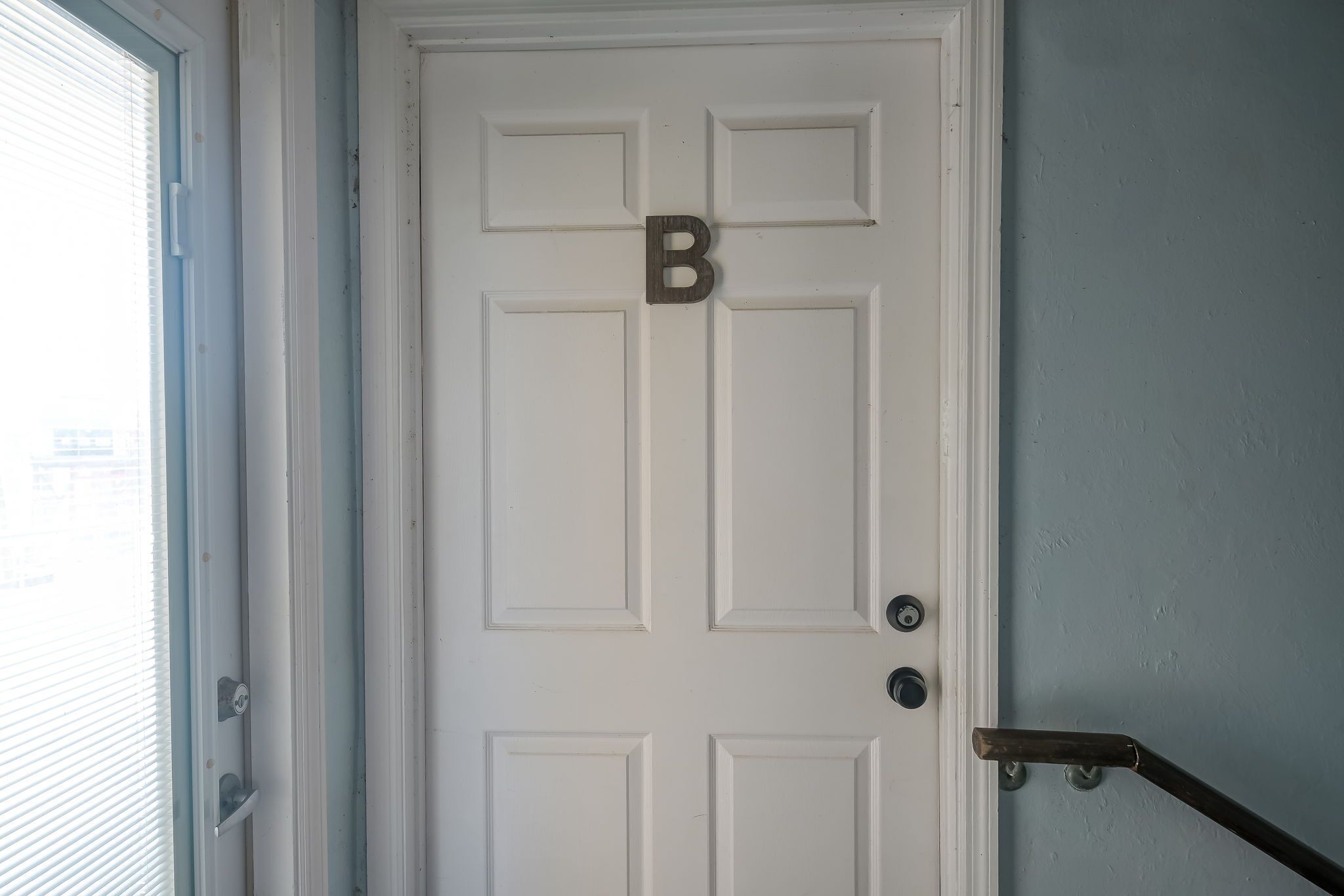 Una puerta blanca con un cartel grande con la letra 'B', un picaporte y un tirador de metal a la derecha.