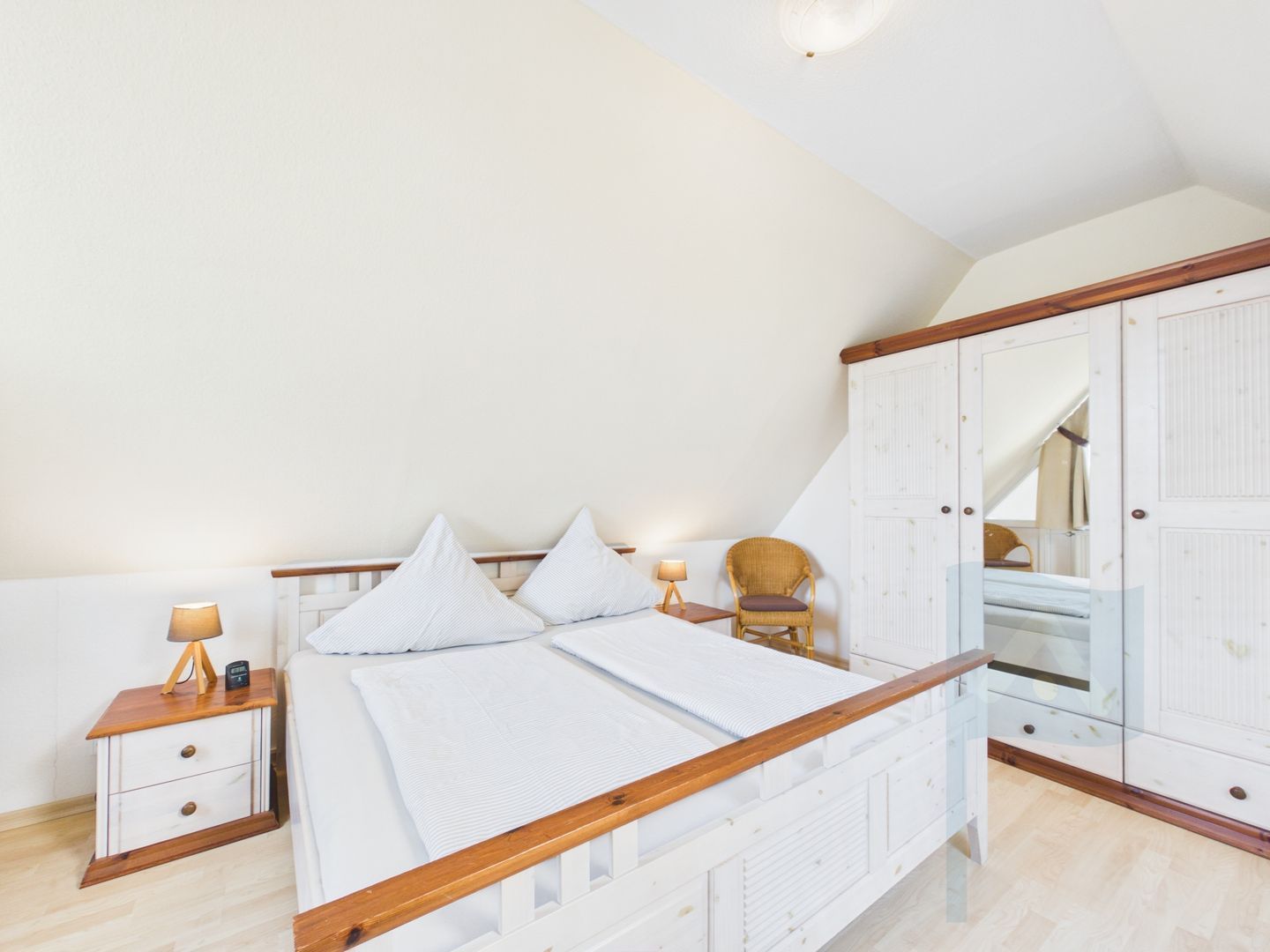 A bedroom with a wooden bed frame, white bedding, a nightstand with a lamp, a chair, and a wardrobe.
