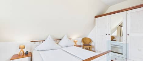 A bedroom with a wooden bed frame, white bedding, a nightstand with a lamp, a chair, and a wardrobe.