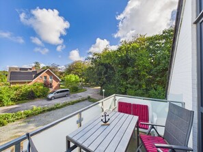 A balcony with a wooden table and red chairs, overlooking a street with a parked car and a house with solar panels.