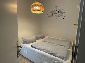 Un dormitorio con cama, dos almohadas, una mesita de noche, un cajón y una lámpara fijada a la pared.