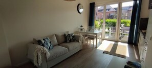 Ein Wohnzimmer mit einer Couch, einem Esstisch und Stühlen. Eine Glastür führt zu einem Außenbereich mit Blick auf Gebäude und Grünflächen.