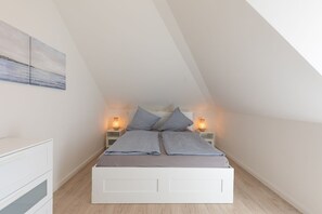Ein Schlafzimmer mit schrägverdeckter Decke, einem Bett mit zwei Kissen, Nachttischlampen und einer weißen Kommode.