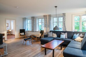 A modern living room with a grey sofa, a wooden coffee table, and a dining area with a small table and chairs. Large windows with patterned curtains allow natural light to fill the space. - Alte Schule Oevenum - Old School in Oevenum (Oevenum)