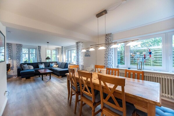 A spacious living room with a dark-colored sofa, a wooden dining table with chairs, and large windows offering a view of the outdoors.