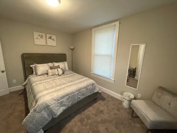 Une chambre à coucher comprenant un lit, une table de chevet, une chaise, une fenêtre avec des stores et un miroir fixé au mur.
