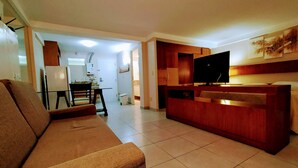 Een woonkamer met een bank, een televisie en een houten kast. Een deur leidt naar een badkamer en in de achtergrond is een keuken te zien.