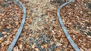 Ein kreisförmiger Sitzbereich im Freien mit Adirondack-Stühlen um eine zentrale Feuerstelle, umgeben von einem Kiesweg und einem bewaldeten Bereich.