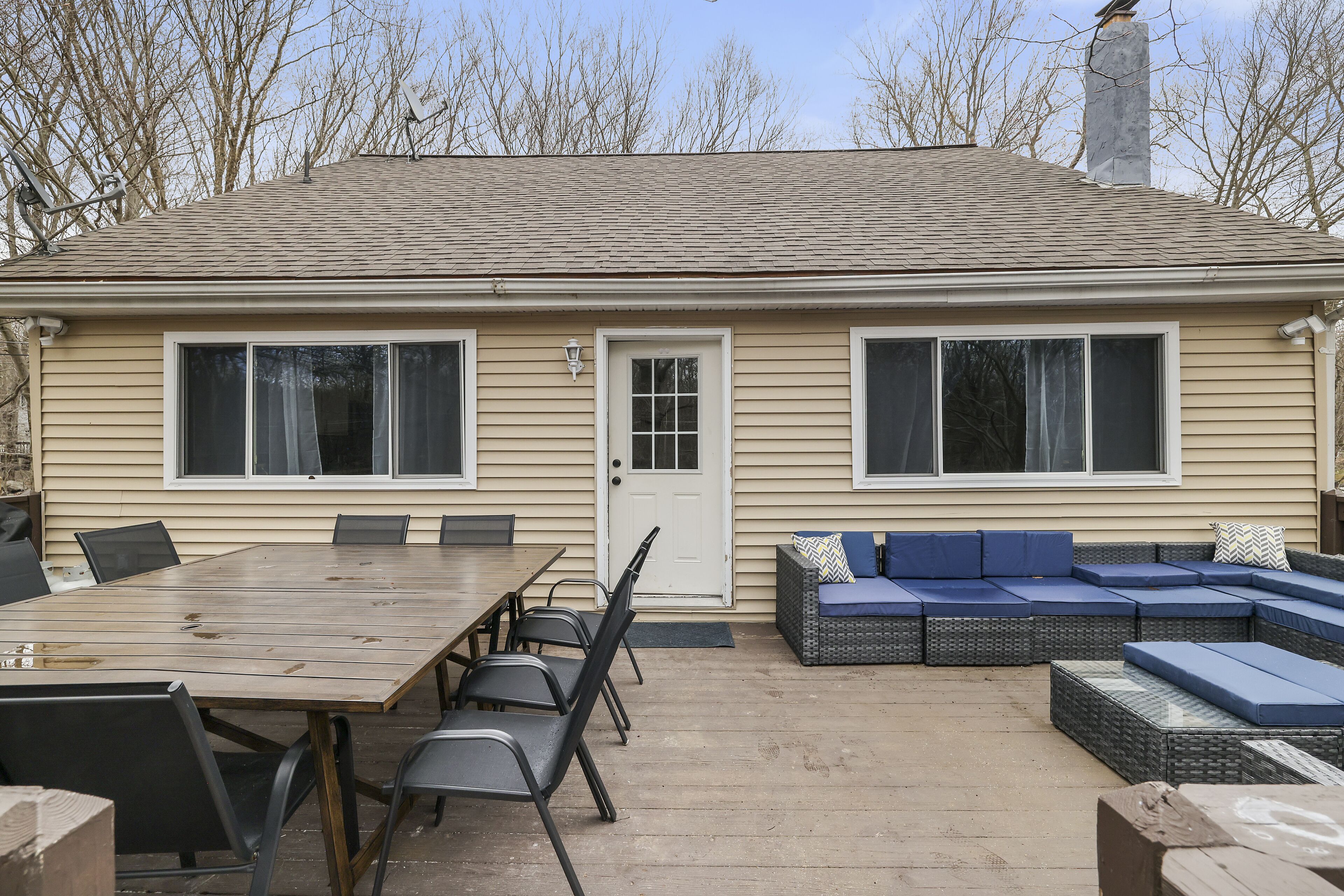 A house with a large patio area featuring a wooden table and chairs, a wicker sofa set, and a door with a window.
