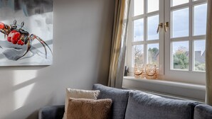 Una sala de estar con un sofá gris, dos cojines, un cuadro de un insecto rojo, y una ventana con cortinas y dos jarrones de cristal.