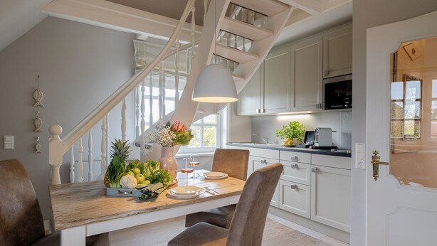 Una cocina moderna con armarios blancos, una mesa de comedor de madera con sillas marrones y una escalera con barandilla blanca.