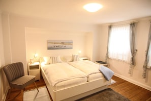 Un dormitorio con cama, mesitas de noche, una silla, un cuadro en la pared y una ventana con cortinas.