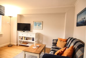 Un salon avec un téléviseur posé sur un meuble blanc, un canapé avec des coussins à carreaux et un tableau accroché au mur.