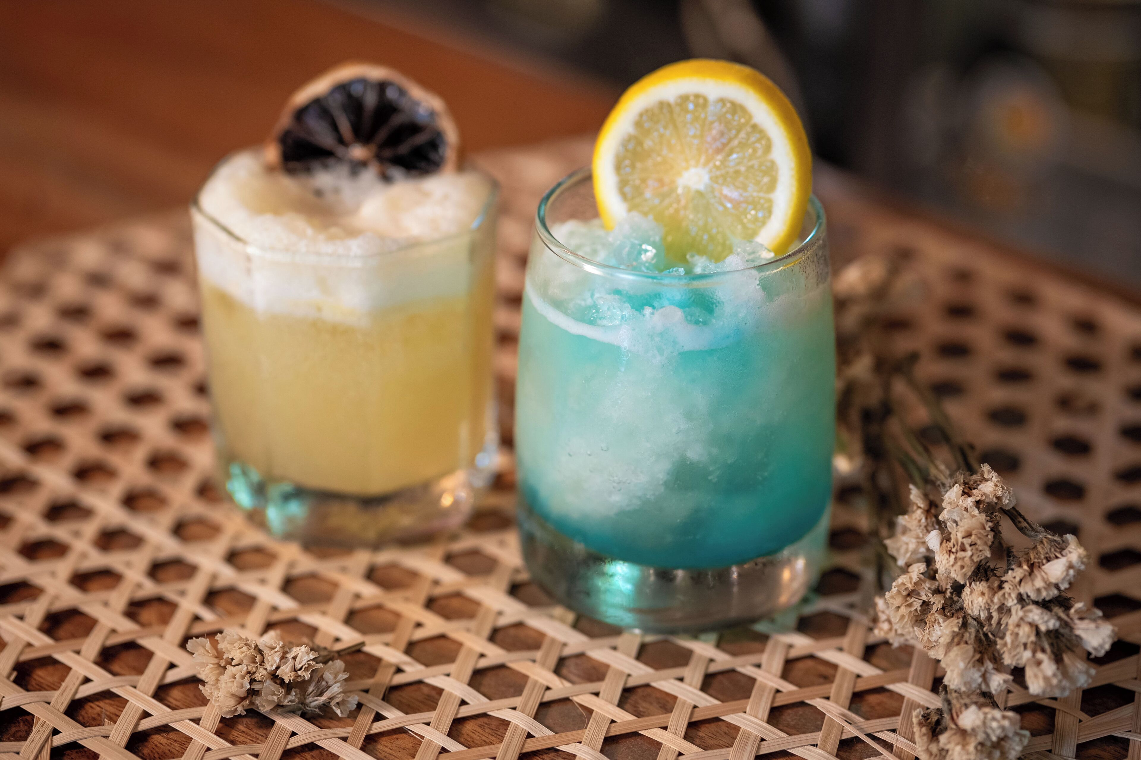 Two glasses of iced drinks on a woven table with dried flowers.