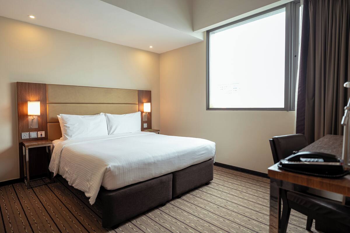 A hotel room with a large bed, two bedside lamps, a wooden headboard, a desk with a telephone, and a large window with curtains.