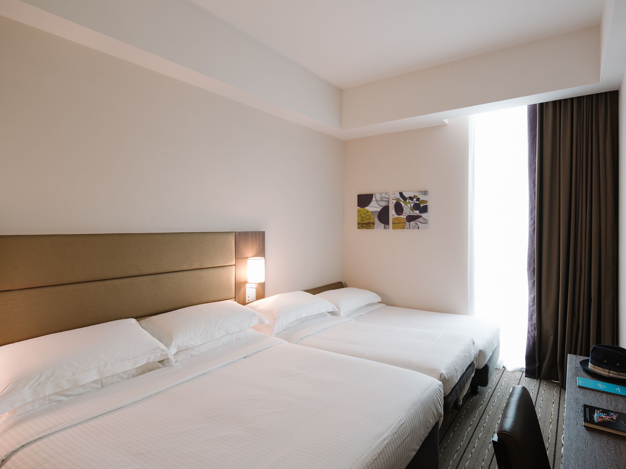 A hotel room with two beds, a nightstand, a lamp, a painting, a window with curtains, and a hat on the table.