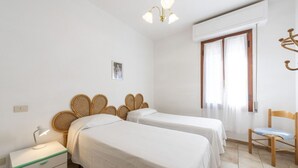 Une chambre avec deux lits simples, une table de chevet avec une lampe, une chaise en bois et une fenêtre avec des rideaux.