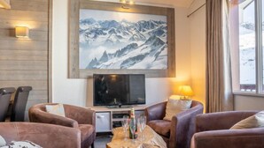 A cozy living room with leather sofas, a wooden coffee table, a flat-screen TV, and a large mountain painting on the wall.