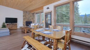 Amplia sala de estar con una mesa de comedor de madera dispuesta para cuatro personas, chimenea de piedra, televisor grande y un rincón de descanso acogedor.