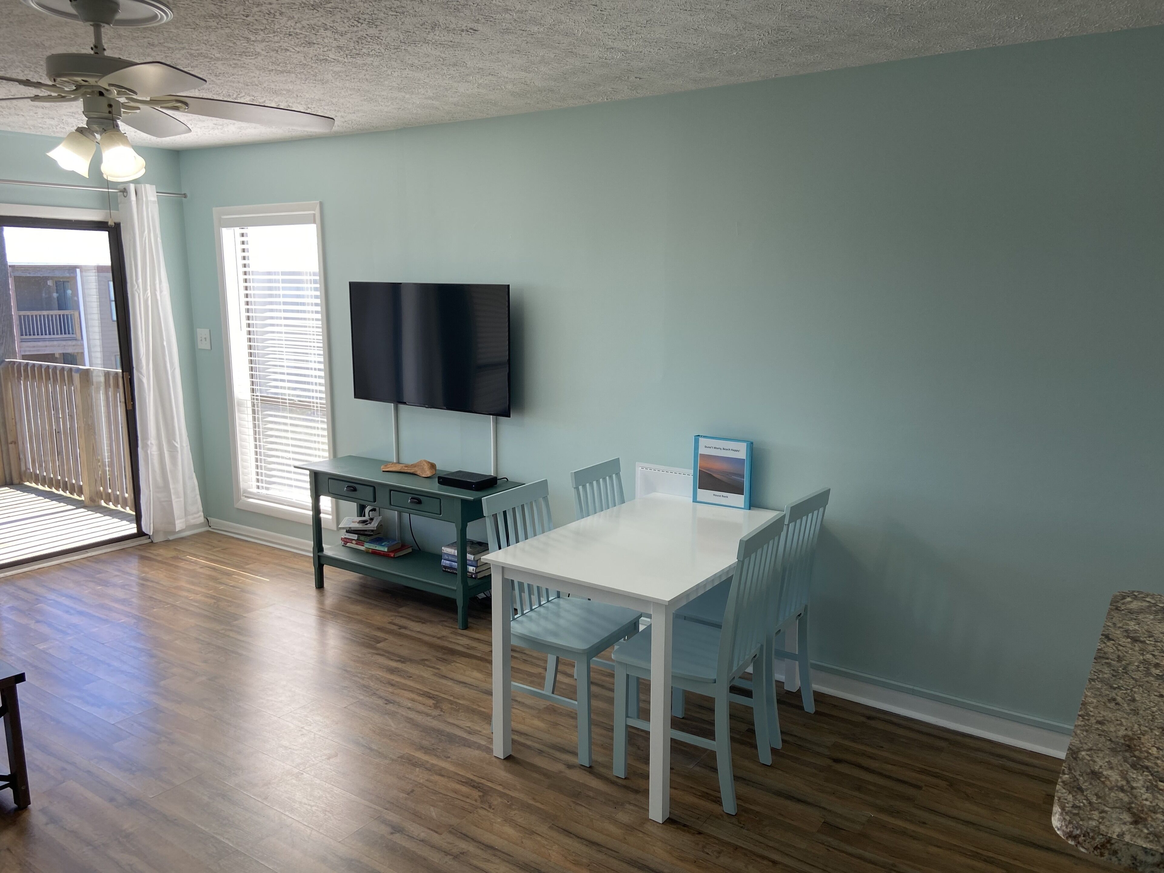 A dining area with a white table and blue chairs, a television mounted on the wall, and a window with blinds.