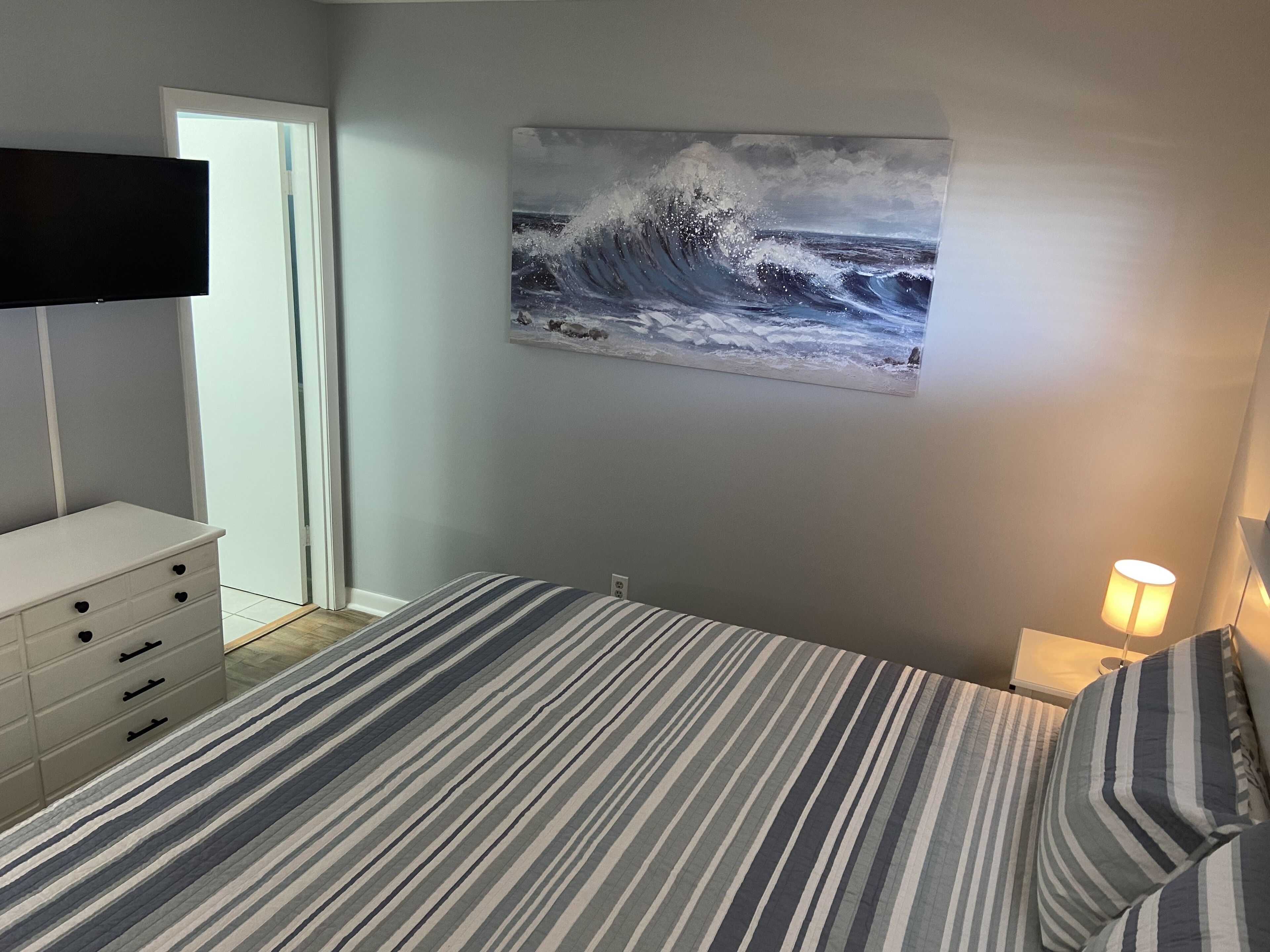 Ett sovrum med en säng, en byrå, en tavla som föreställer en våg och en lampa.