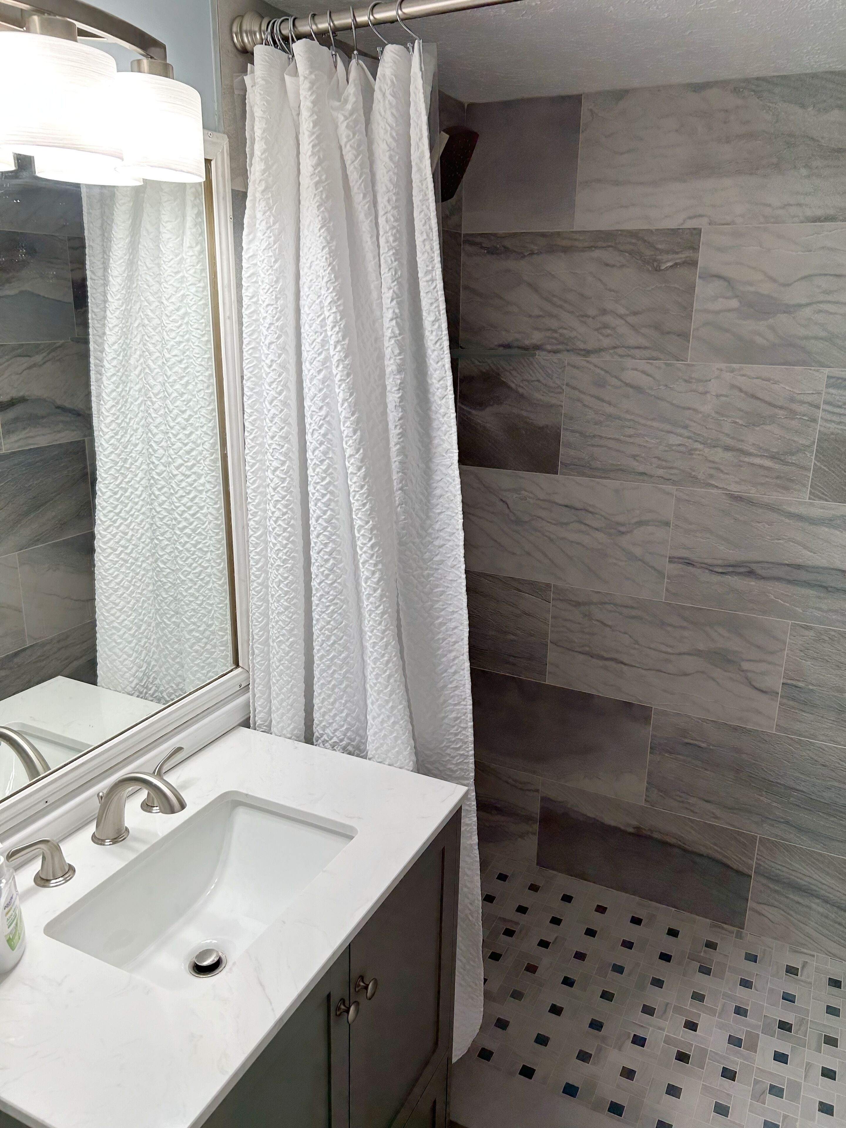 A bathroom with a white sink, a mirror, a shower curtain, and a tiled shower wall.