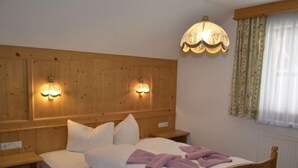 Een houten bed met witte beddengoed en paarse handdoeken, twee wandlampen en een raam met bloemen gordijnen.