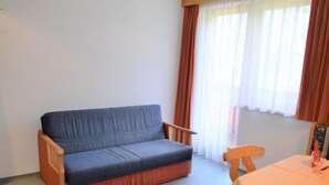 Een kleine kamer met een blauwe bank, een houten stoel, een kleintje tafel met een witte tafelkleed en een raam met dunne gordijnen.