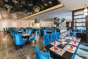 Uma ampla área de jantar com pé-direito alto e iluminação moderna, com cadeiras e mesas azuis dispostas para refeições.