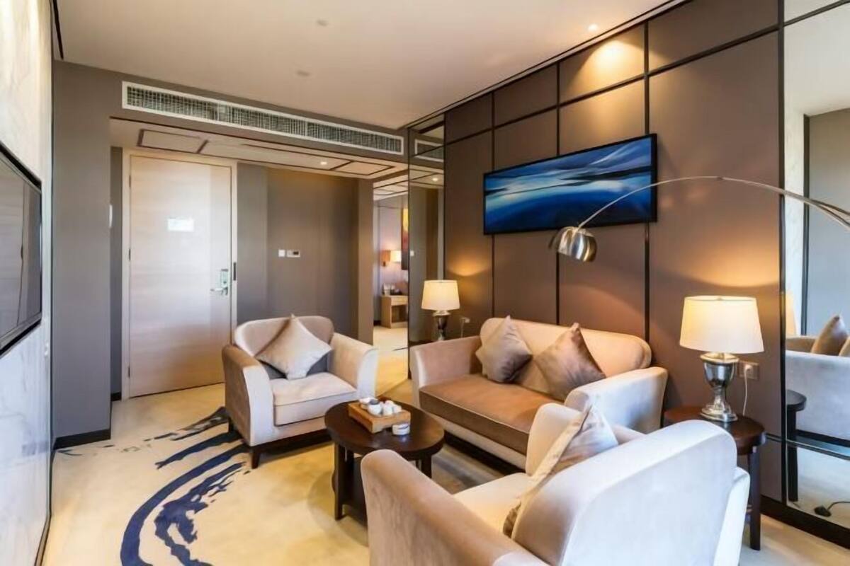 A modern hotel room with a sofa, armchairs, a coffee table, and a TV mounted on the wall.