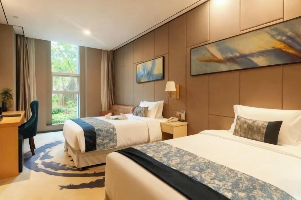 A hotel room with two beds, a desk, a chair, a window with curtains, and artwork on the walls.