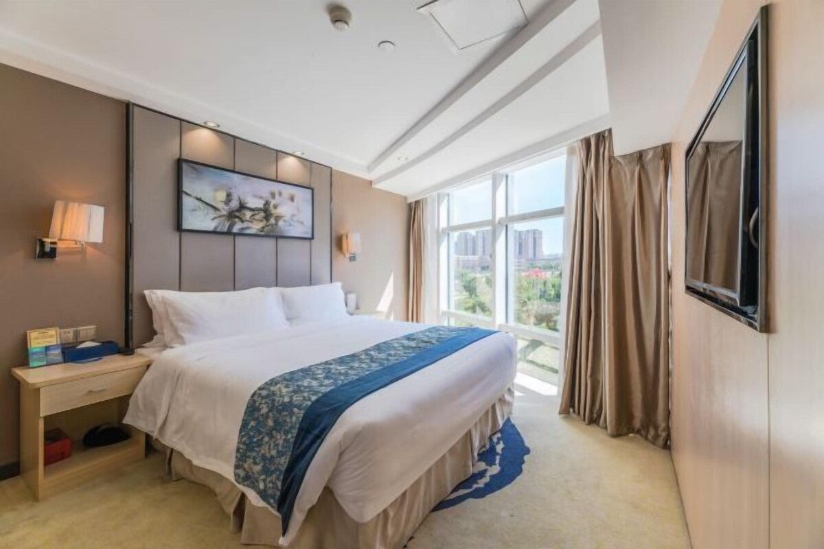 Habitación de hotel con una cama grande, una mesita de noche, un televisor instalado en la pared y una ventana con cortinas.