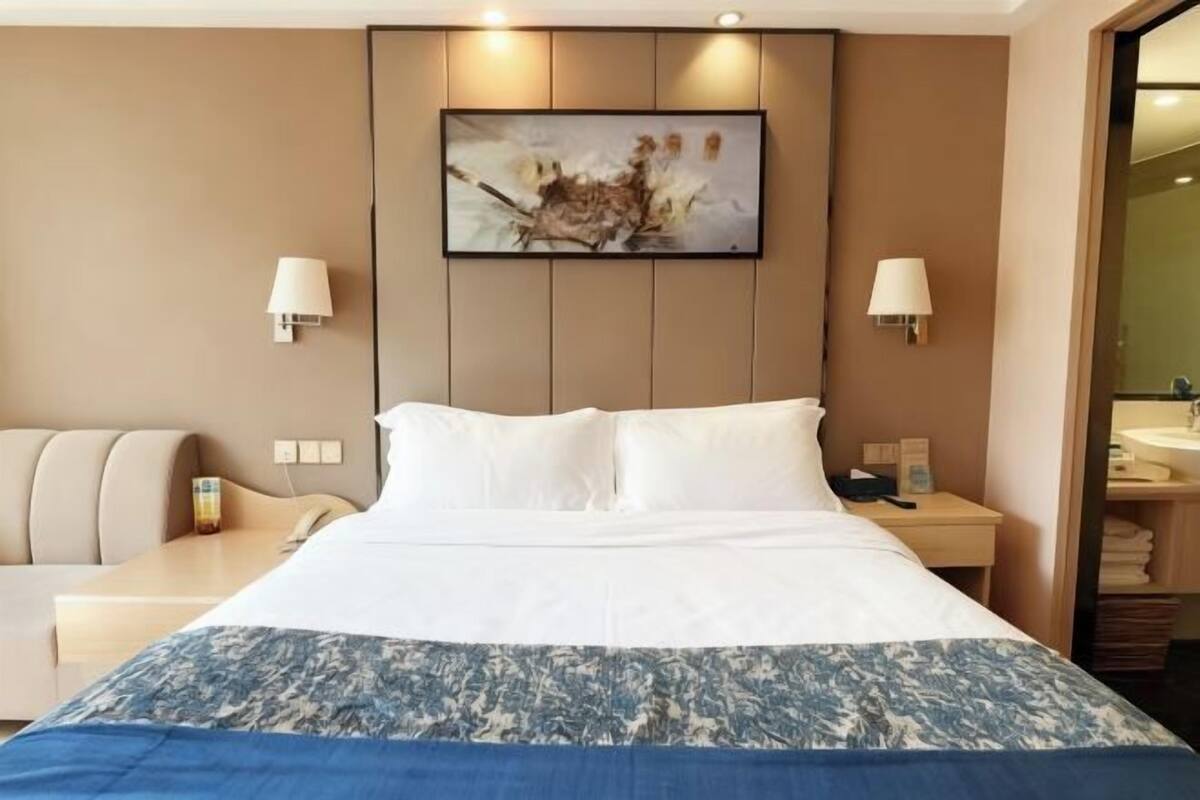 A hotel room with a large bed, two bedside tables, a desk, a chair, and a bathroom visible through an open door.