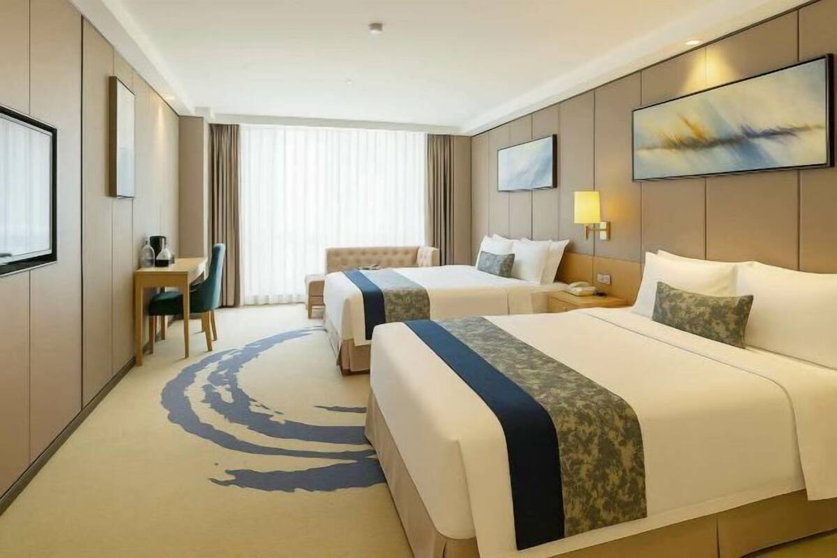 A hotel room with two beds, a desk, a chair, a large window, and artwork on the walls.