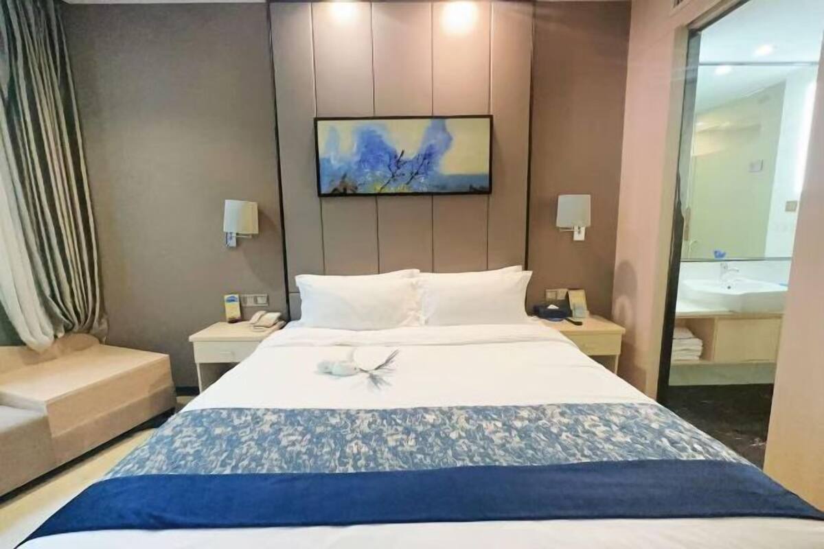 A hotel room with a bed, bedside tables, a painting, a mirror, and a bathroom visible through a glass door.