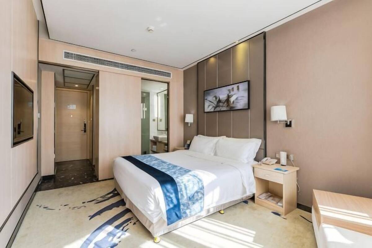 A hotel room with a bed, bedside table, lamp, and a painting on the wall.
