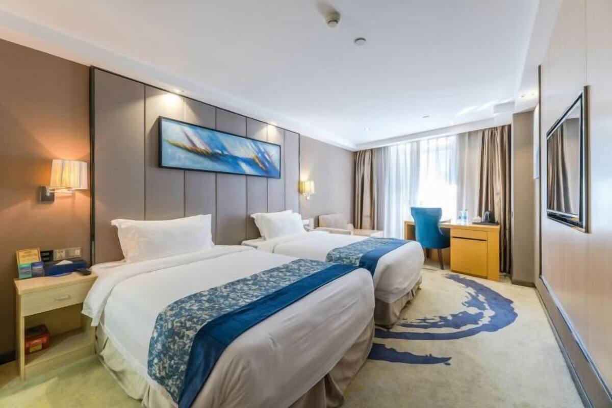 A hotel room with two beds, a desk, a chair, a painting on the wall, and a large window with curtains.