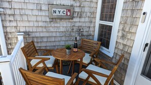 A wooden deck with a table and chairs, a potted plant, and a bottle of wine. A sign with "NYC" is visible on the wall. - Be right in the action in our centrally-located 2BR/1BA condo with Hot Tub (Provincetown)