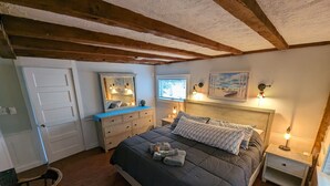 A bedroom with a large bed, wooden beams on the ceiling, a dresser, a mirror, and a window with blinds. - Be right in the action in our centrally-located 2BR/1BA condo with Hot Tub (Provincetown)