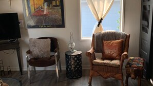 Una sala de estar acogedora con un cuadro grande de un café, una silla de mimbre, una mesita auxiliar y vistas al exterior a través de una ventana.