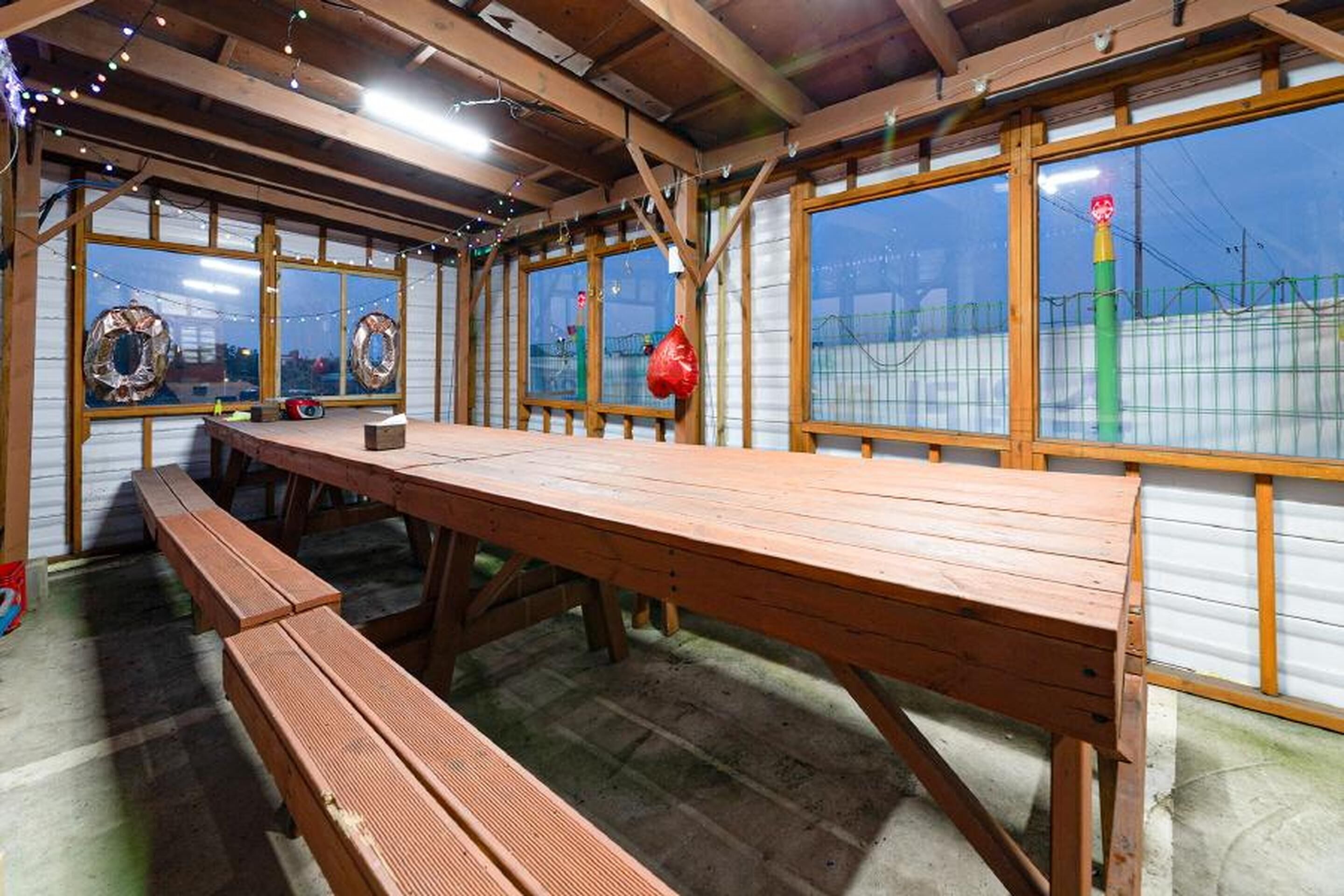 Une table et des bancs en bois se trouvent dans une pièce au décor rustique, dotée de grandes fenêtres et offrant une vue sur un espace clos.