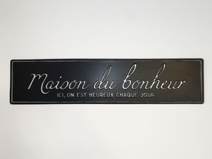 A black sign with white cursive text that reads "Maison du bonheur" and "ICI, ON EST HEUREUX CHAQUE JOUR".
