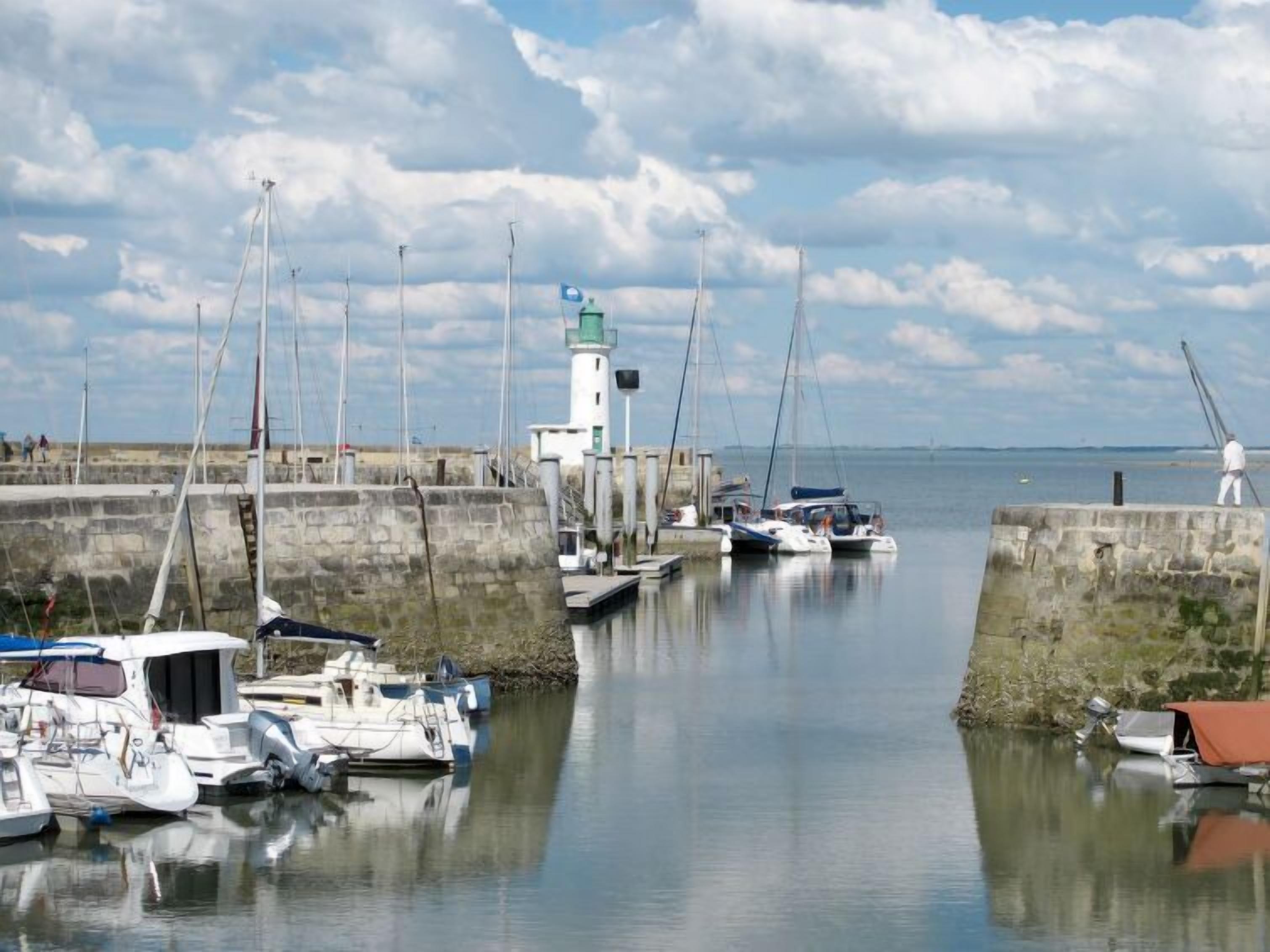 Un port où sont amarrés des bateaux, un phare et une mer calme sous un ciel partiellement nuageux.
